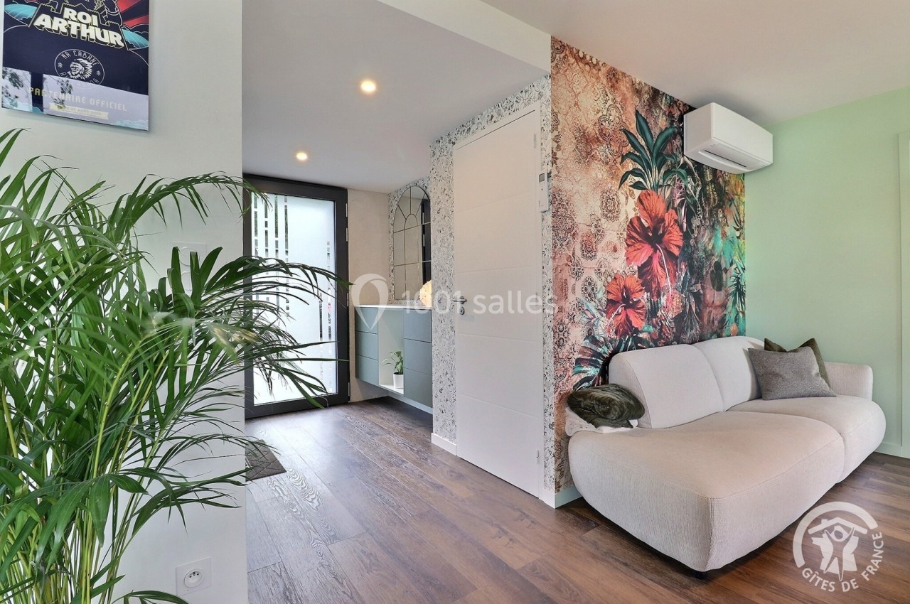 Salon lumineux avec canapé beige, mur décoré de motifs floraux, sol en parquet et vue sur une entrée moderne.