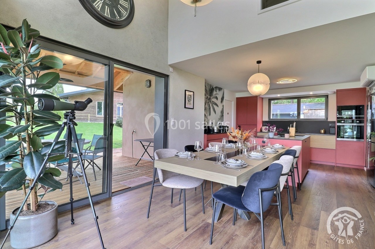 Salle à manger moderne avec table dressée, cuisine ouverte en arrière-plan et accès à une terrasse extérieure.