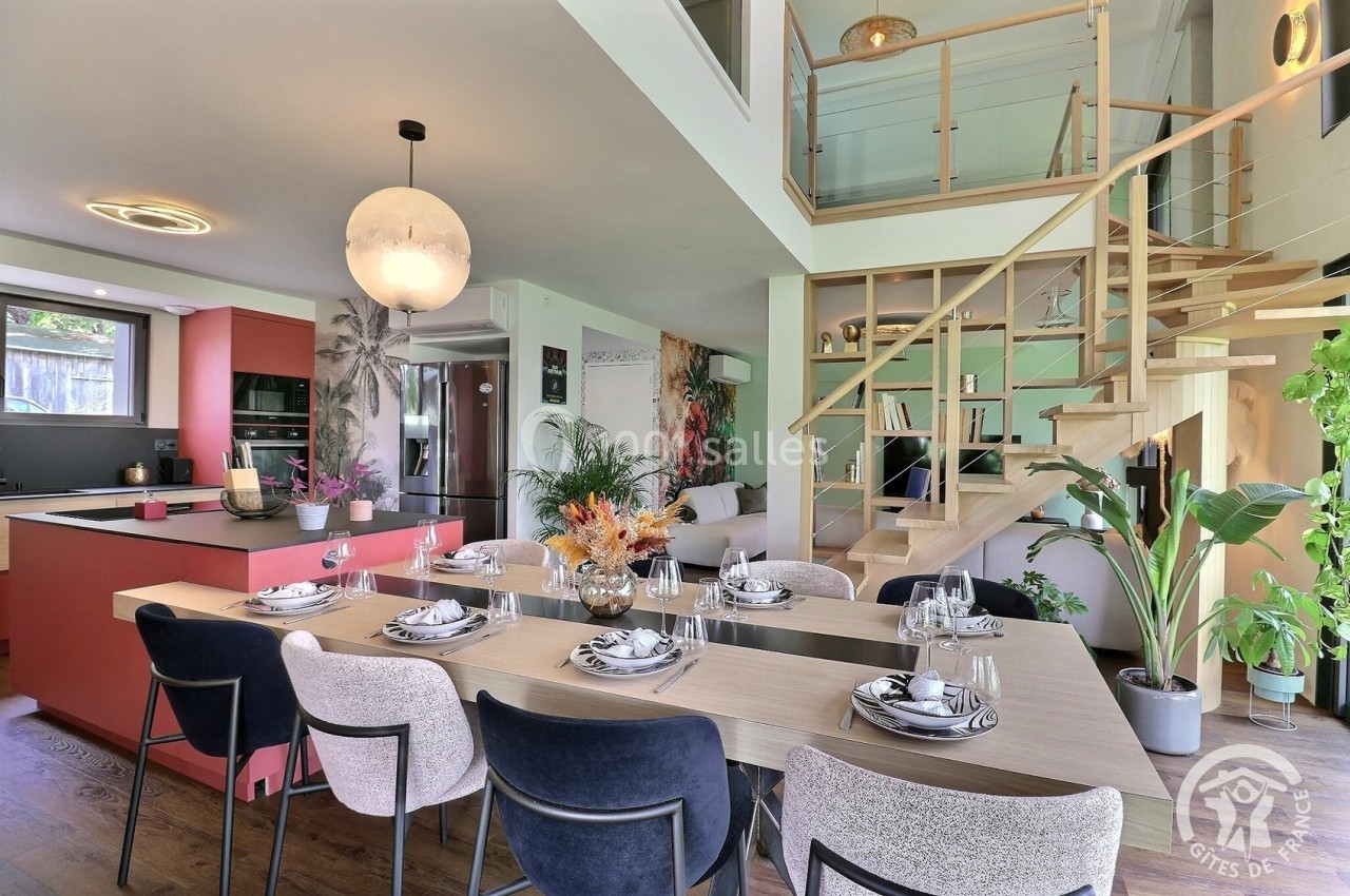 Salle à manger moderne avec table dressée, cuisine ouverte en arrière-plan et escalier menant à un étage.
