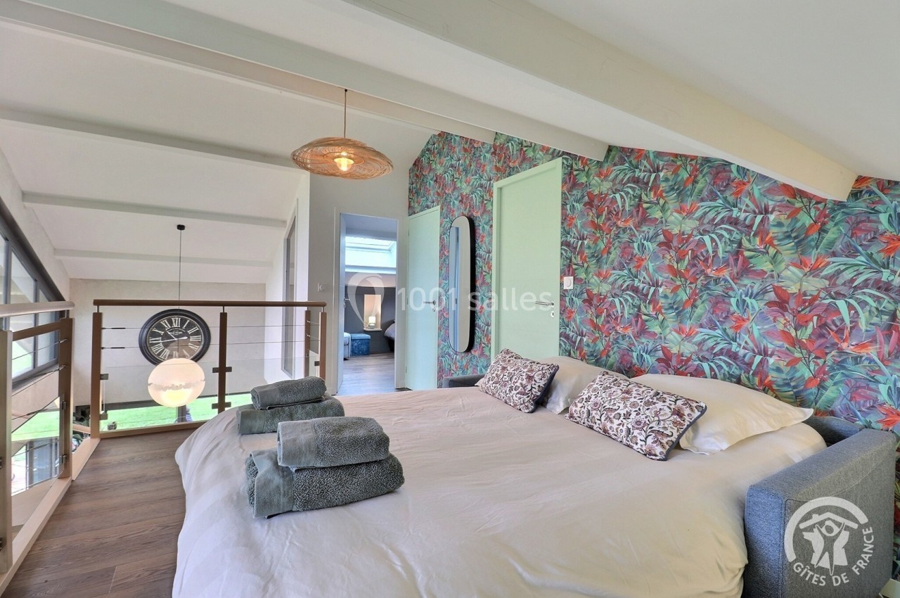 Chambre lumineuse avec lit double, linge plié, mur à motifs tropicaux et vue sur mezzanine avec horloge murale.