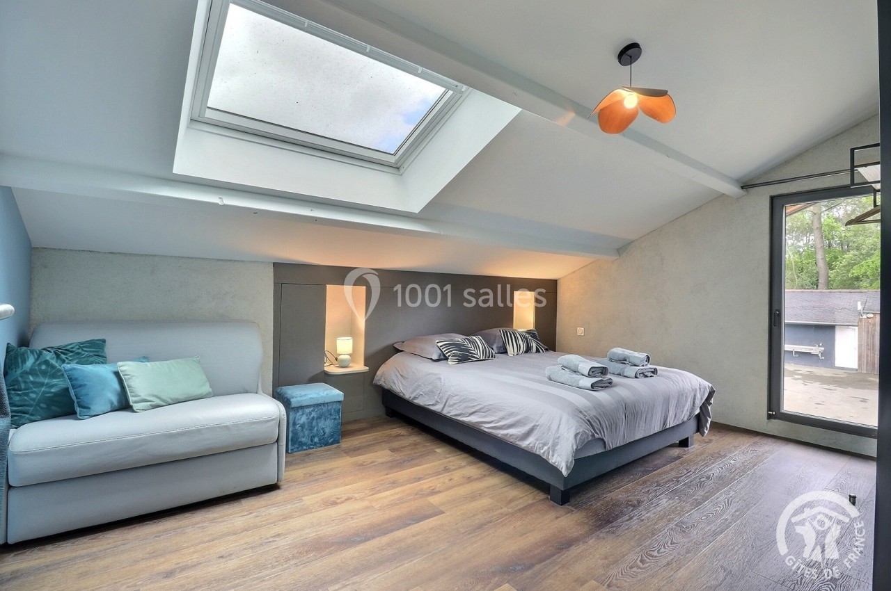 Chambre lumineuse sous les combles avec lit double, canapé, parquet, grande fenêtre et puits de lumière.