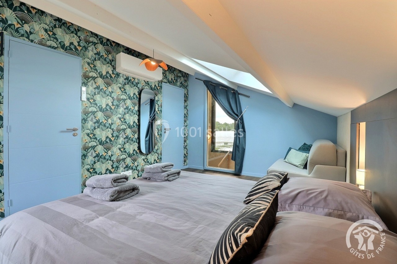 Chambre mansardée avec lit double, décoration murale végétale, fauteuil, fenêtre et serviettes pliées sur le lit.