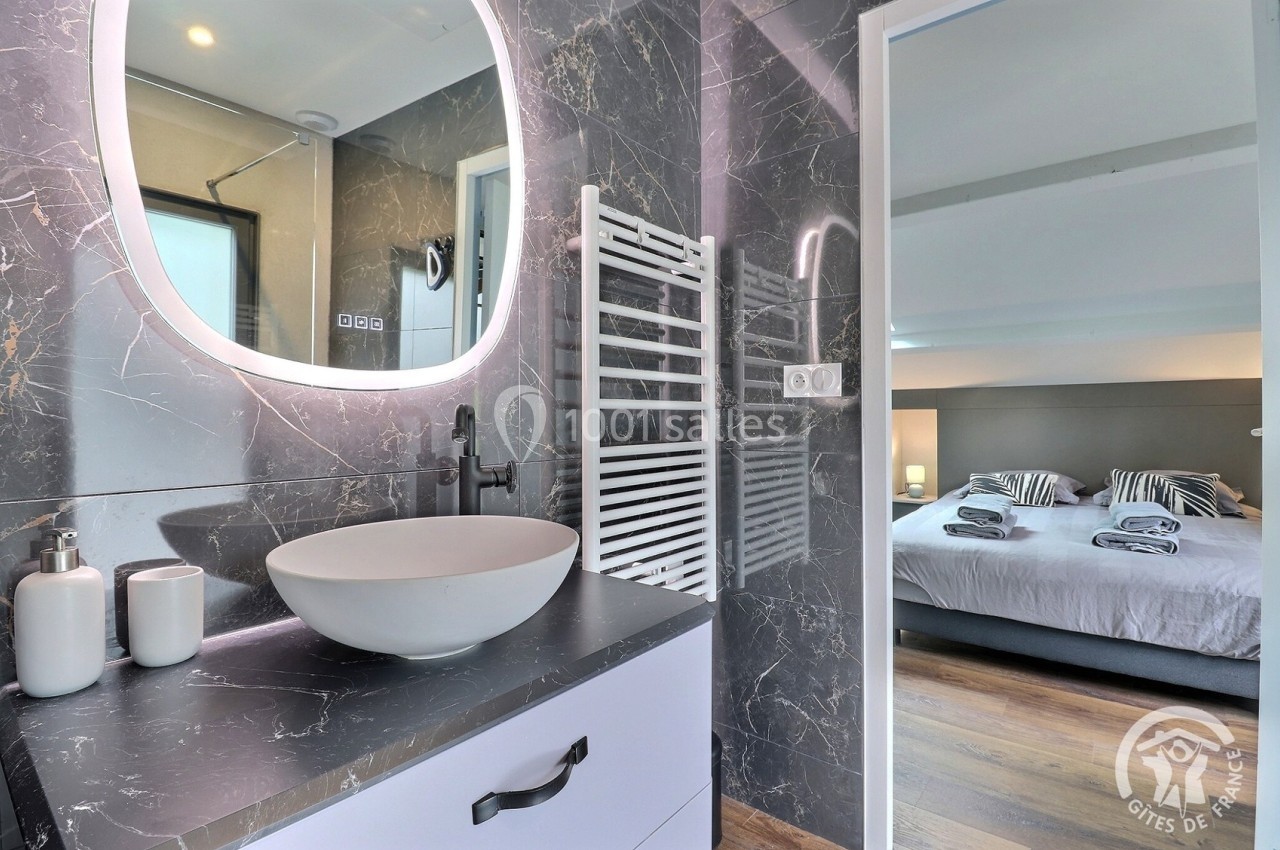 Salle de bain moderne avec vasque blanche, miroir éclairé, et vue sur une chambre avec lit double en arrière-plan.