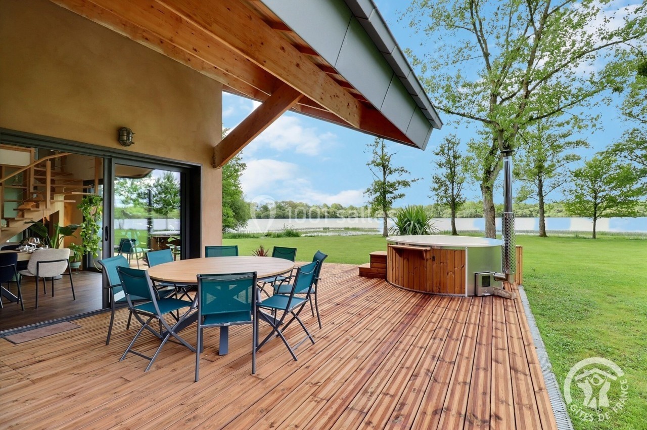 Terrasse en bois avec table, chaises, bain à remous, vue sur un jardin verdoyant et un lac en arrière-plan.