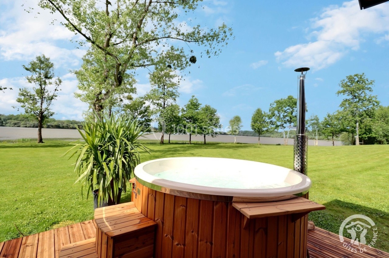 Bain nordique en bois sur une terrasse, entouré de pelouse et d'arbres avec un plan d'eau en arrière-plan.