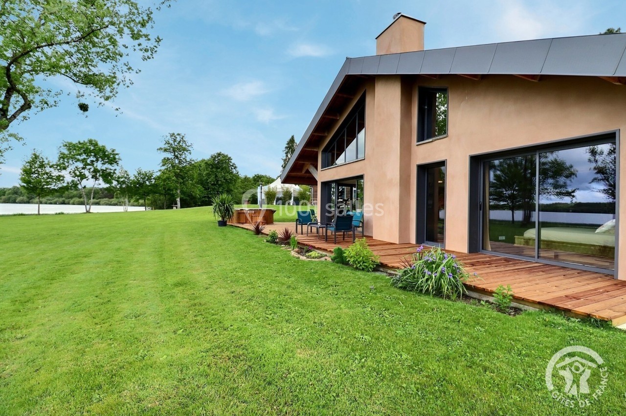 Maison moderne avec terrasse en bois, entourée d'une pelouse et donnant sur un grand espace vert avec un plan d'eau.