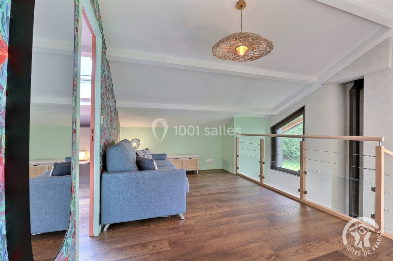 Salon mansardé avec canapé bleu, parquet en bois, rambarde en métal et bois, et lumière naturelle entrant par les fenêtres.