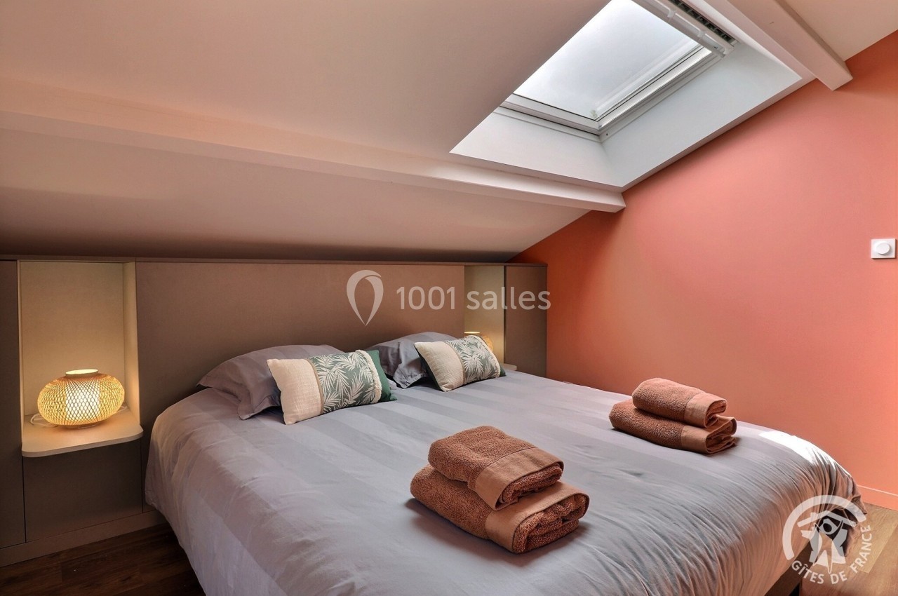 Chambre mansardée avec lit double, linge de lit et serviettes pliées, éclairée par une fenêtre de toit.
