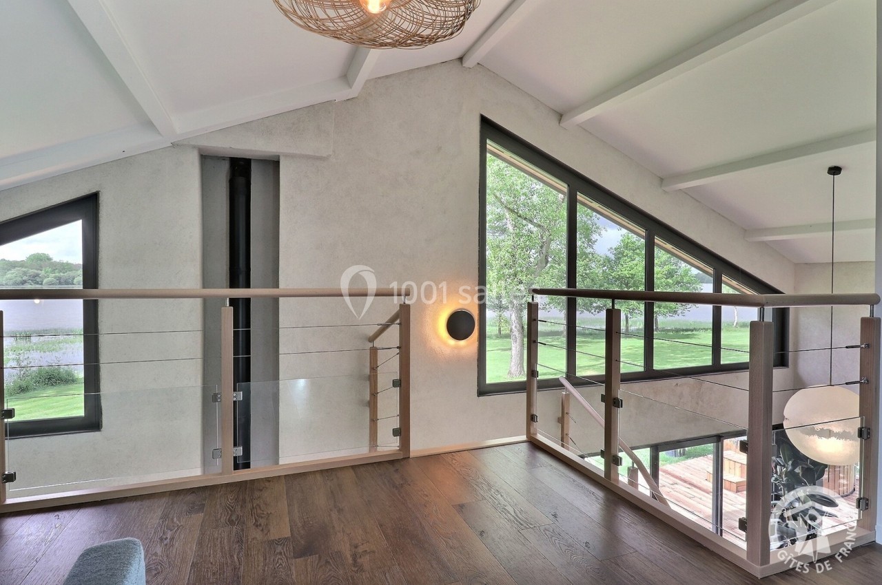 Mezzanine lumineuse avec parquet en bois, garde-corps en verre et vue sur un paysage verdoyant à travers de grandes fenêtres.