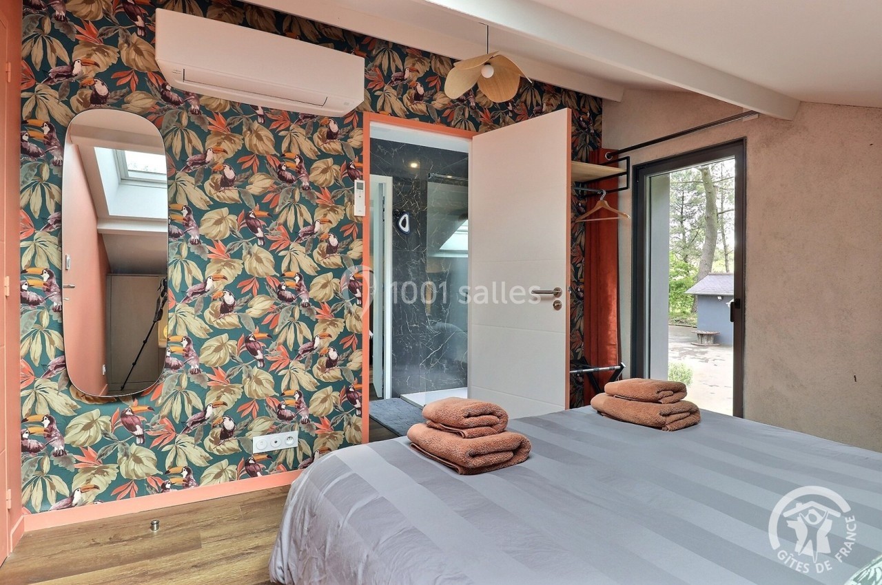 Chambre avec lit double, linge de lit plié, mur à motifs floraux, miroir, climatisation et vue sur une salle de bain.