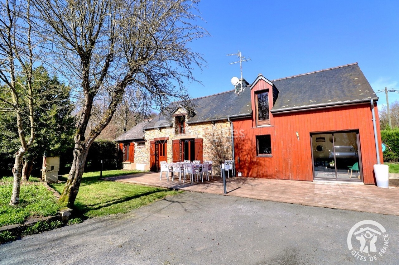 Maison en pierre et bois avec terrasse, table extérieure et jardin, entourée d'arbres sous un ciel bleu.