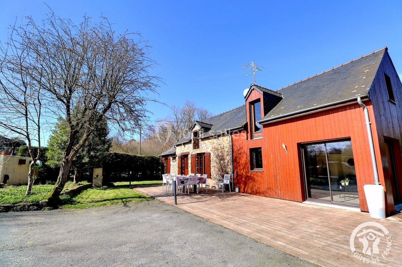 Maison en pierre et bois avec terrasse ensoleillée, table extérieure et jardin arboré, sous un ciel bleu.
