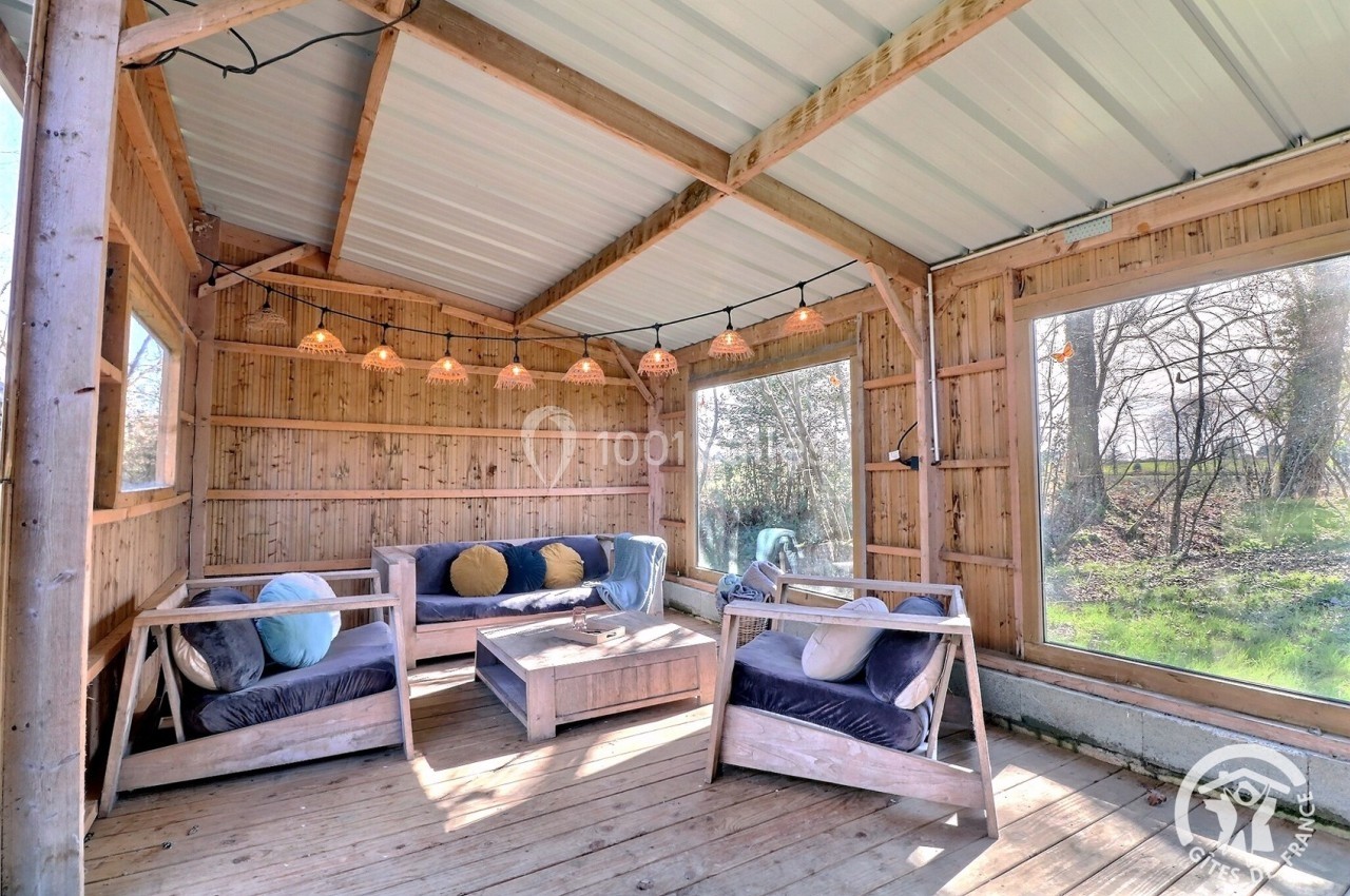 Salon extérieur en bois avec canapés, coussins colorés, guirlande lumineuse et grandes fenêtres donnant sur la nature.