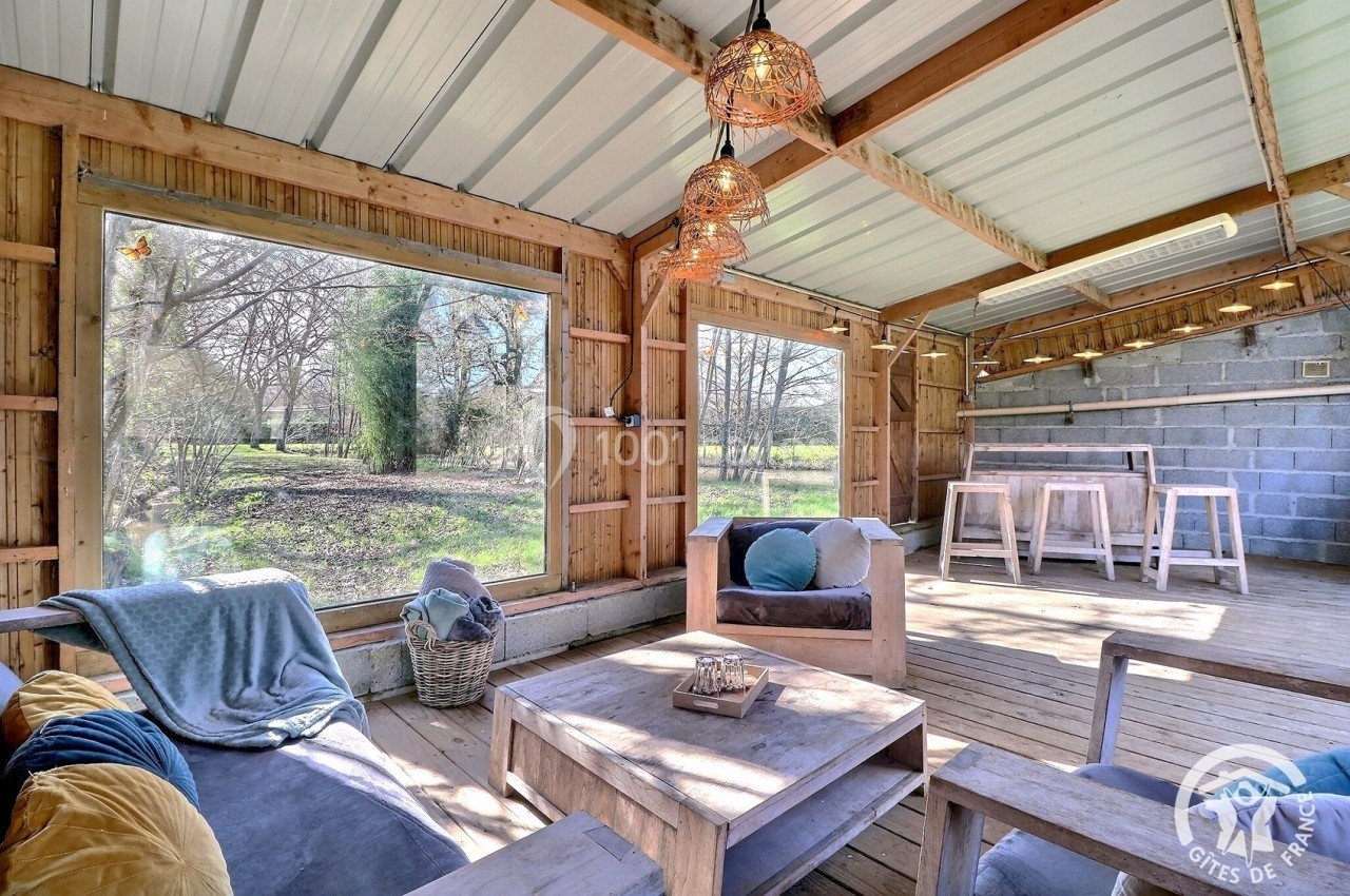 Espace intérieur lumineux en bois avec mobilier rustique, grandes baies vitrées donnant sur un paysage arboré.