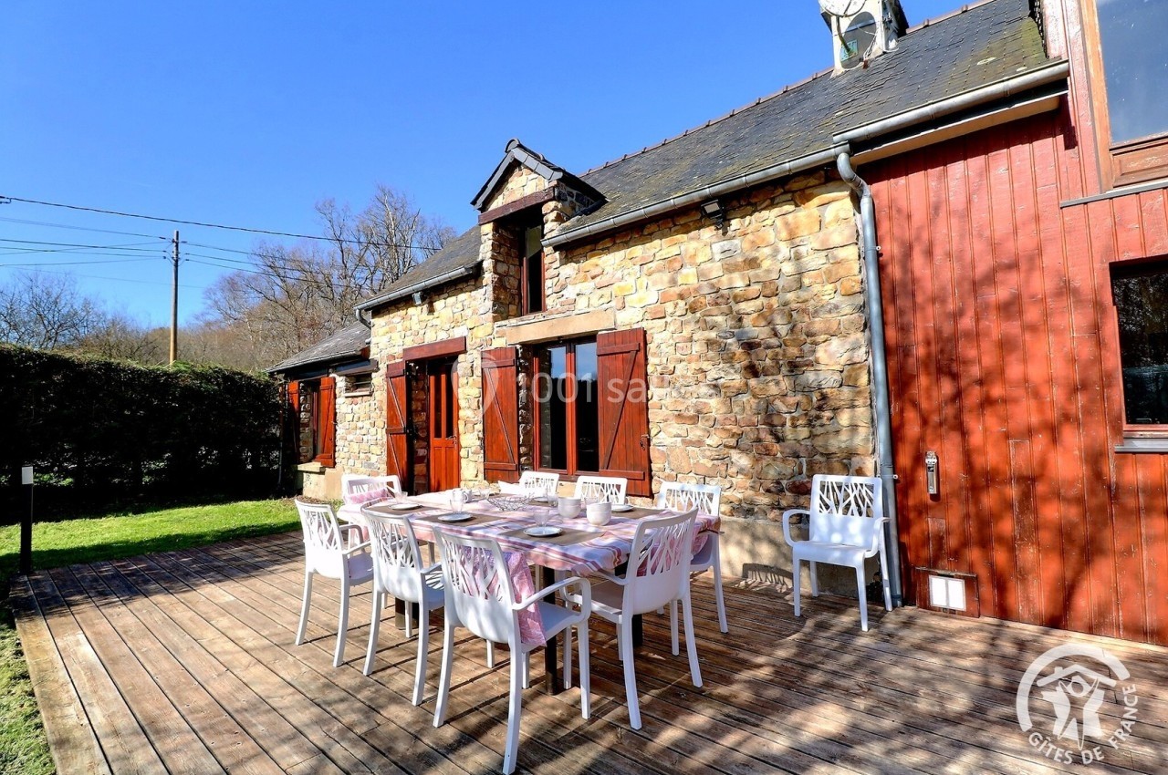 Terrasse en bois avec table dressée et chaises blanches devant une maison en pierre et bois, entourée de verdure.