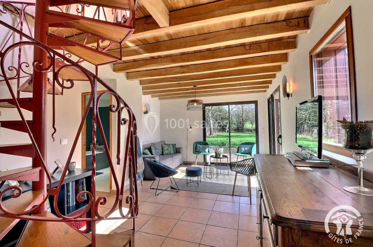 Salon lumineux avec escalier en colimaçon, poutres apparentes, mobilier moderne et vue sur un jardin verdoyant.