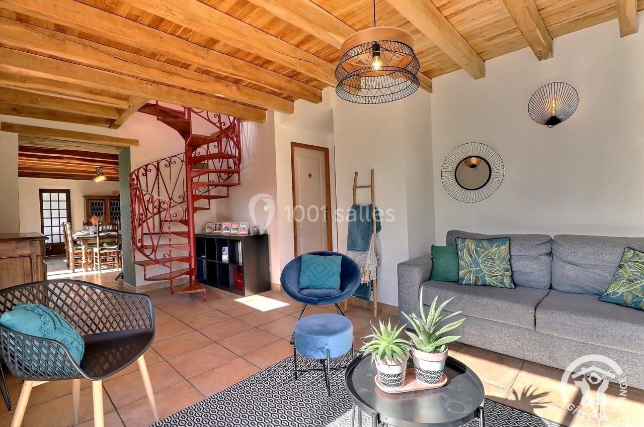 Salon lumineux avec canapé gris, fauteuils, plantes décoratives et escalier en colimaçon en arrière-plan.