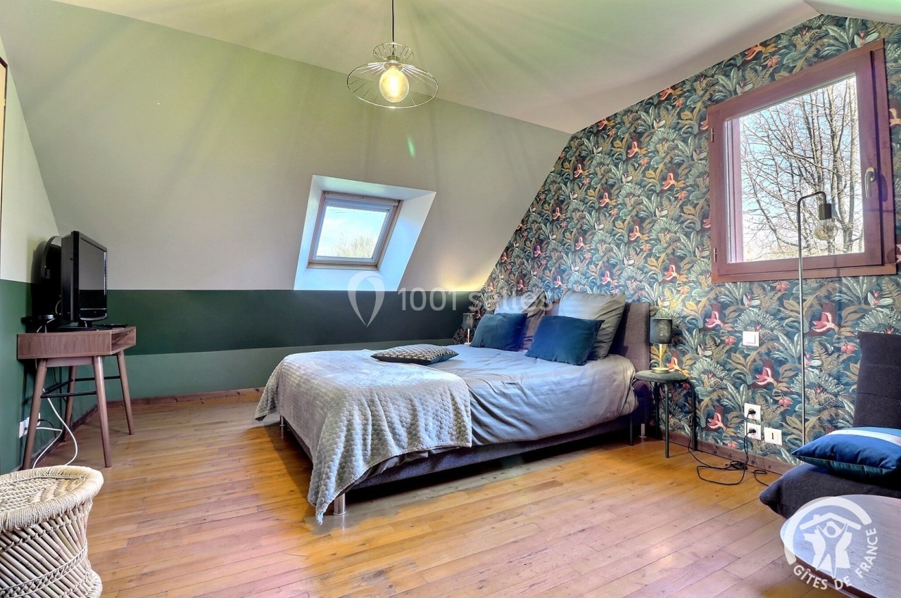 Chambre mansardée avec lit double, parquet en bois, mur tapissé de motifs tropicaux et fenêtre de toit.