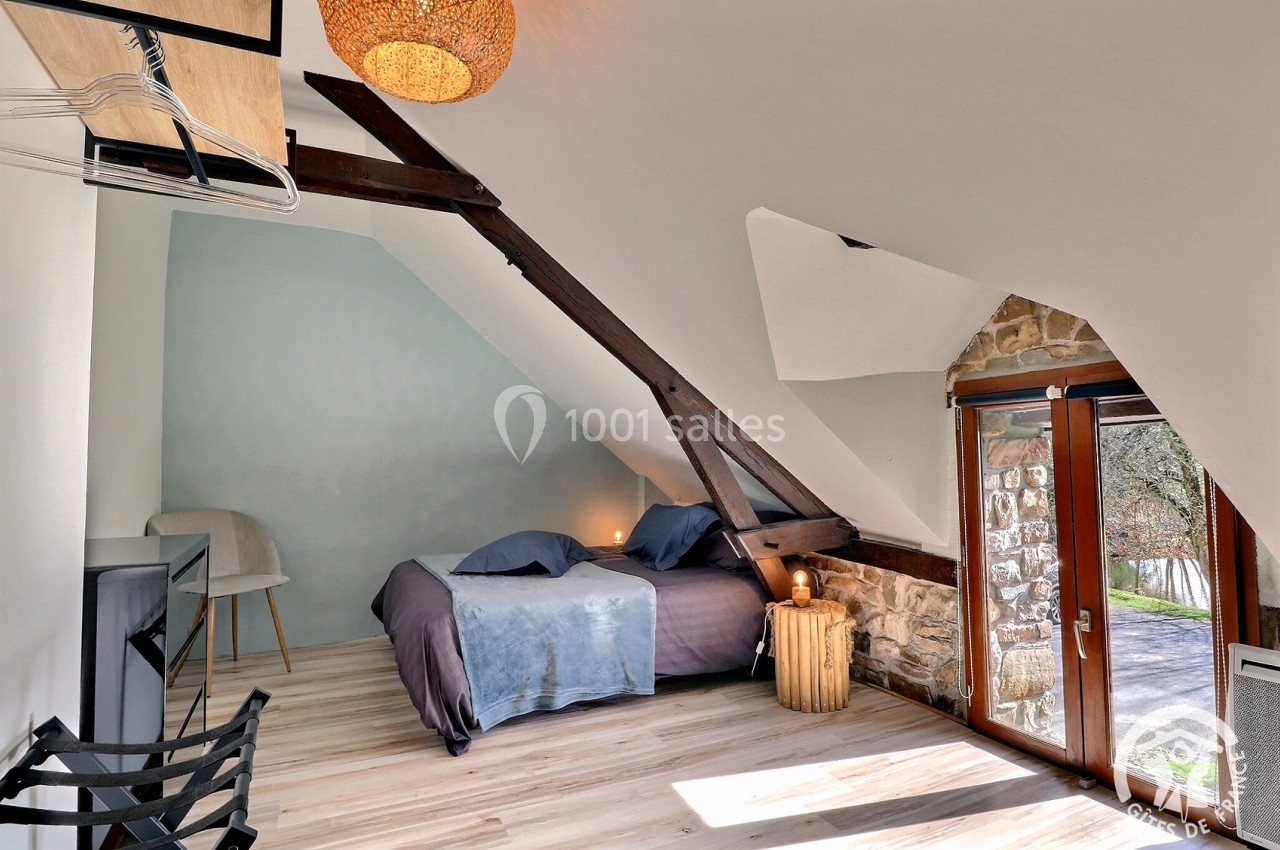 Chambre mansardée avec poutres apparentes, lit double, lumière naturelle et vue sur l'extérieur par une porte vitrée.