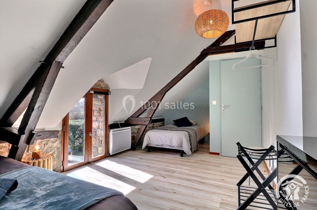 Chambre mansardée avec poutres apparentes, deux lits simples, bureau, chaises pliantes et lumière naturelle.