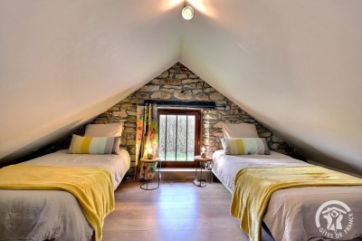 Chambre mansardée avec deux lits simples, murs en pierre apparente, lumière naturelle et décoration chaleureuse.