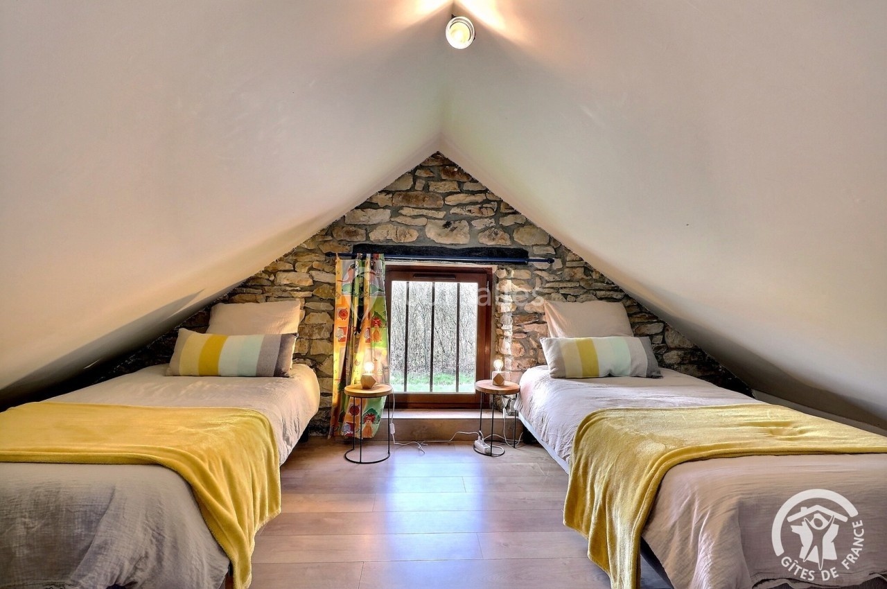 Chambre mansardée avec deux lits simples, murs en pierre apparente, lumière naturelle et décoration chaleureuse.