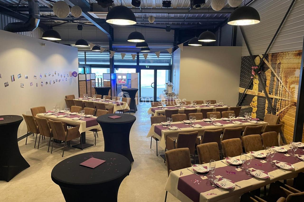 Salle décorée pour un événement avec tables dressées, nappes violettes, chaises rembourrées et éclairage suspendu.