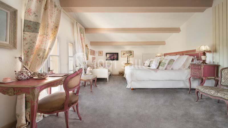 Chambre lumineuse avec lit double, mobilier classique rose et beige, bureau, fauteuils et grandes fenêtres avec rideaux…