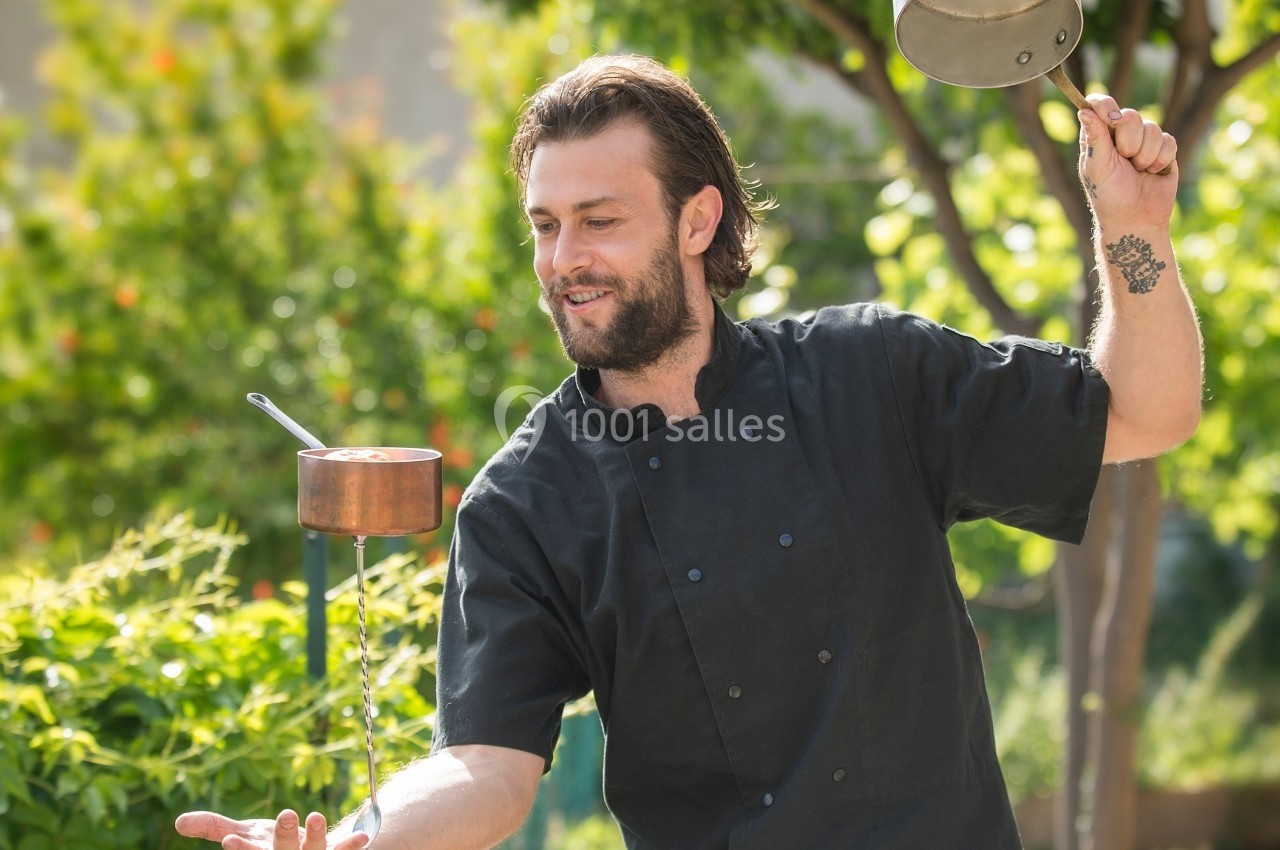 Un chef en tenue noire jongle avec des casseroles en extérieur, entouré de verdure.