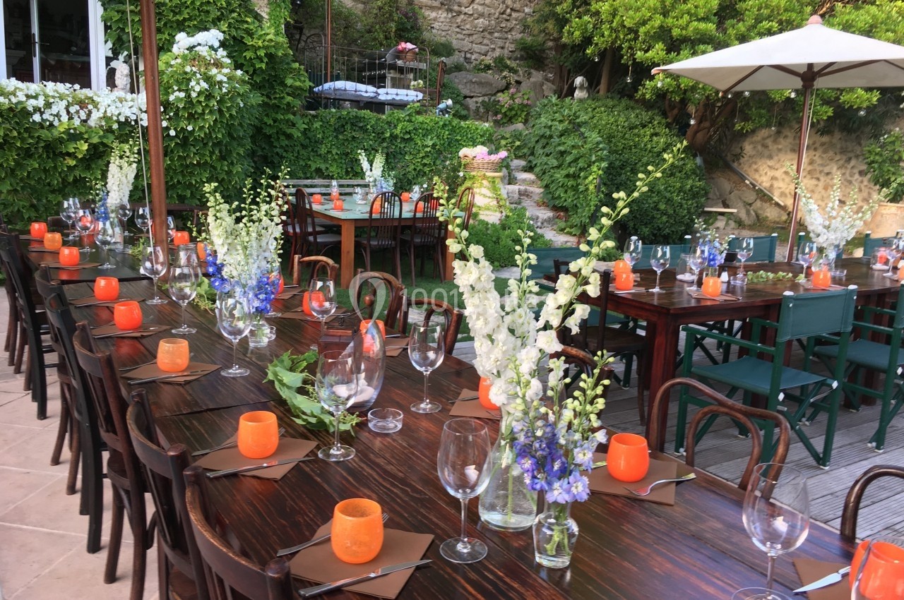 Tables en bois décorées de fleurs blanches et violettes, bougies orange et verres, disposées dans un jardin verdoyant.