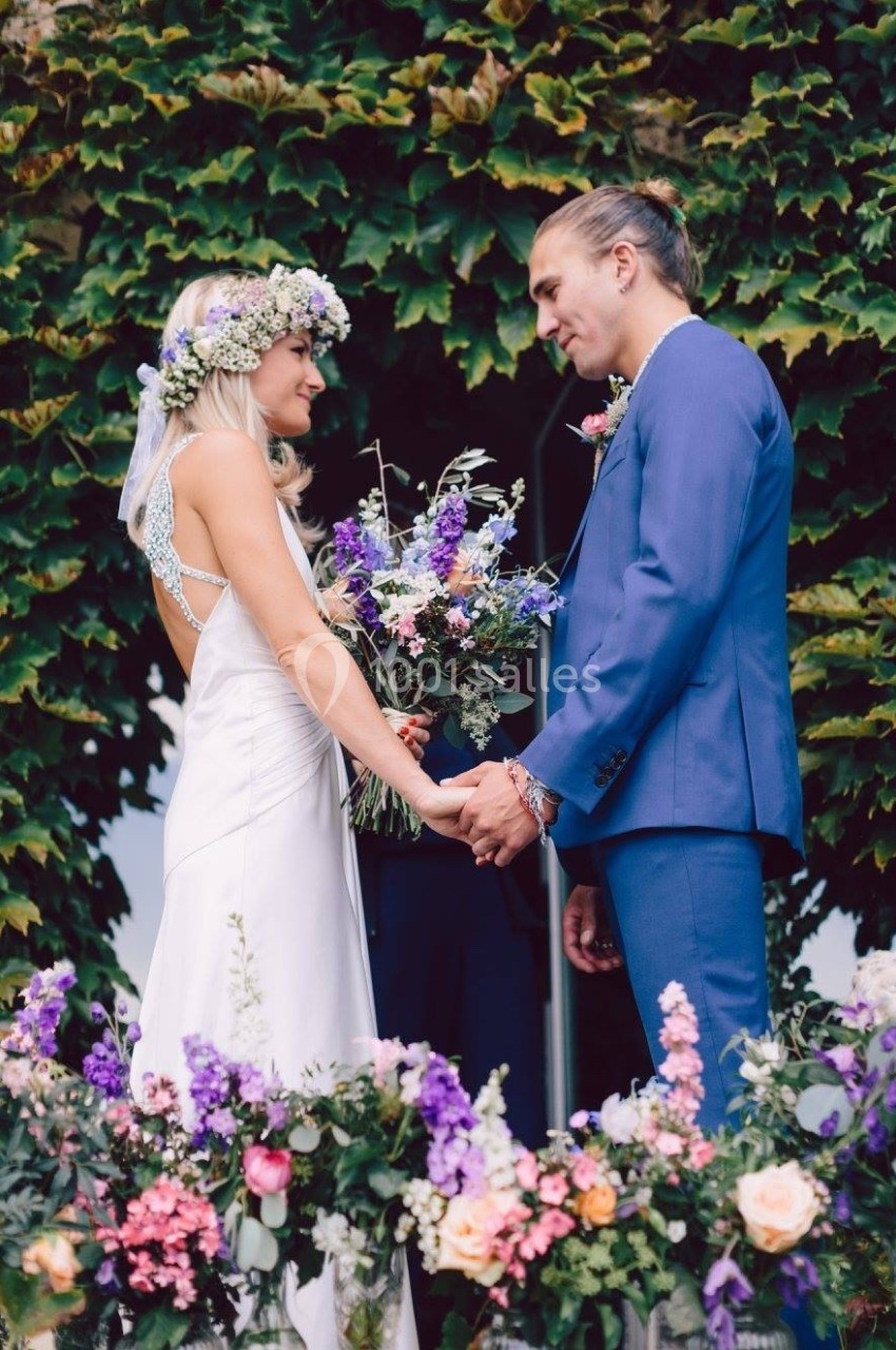 Un couple en tenue de mariage se tient la main devant une arche fleurie, entouré de verdure.