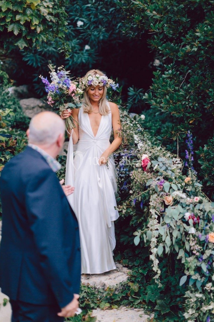 Une femme en robe blanche et couronne de fleurs descend un escalier en pierre, tenant un bouquet, entourée de verdure.