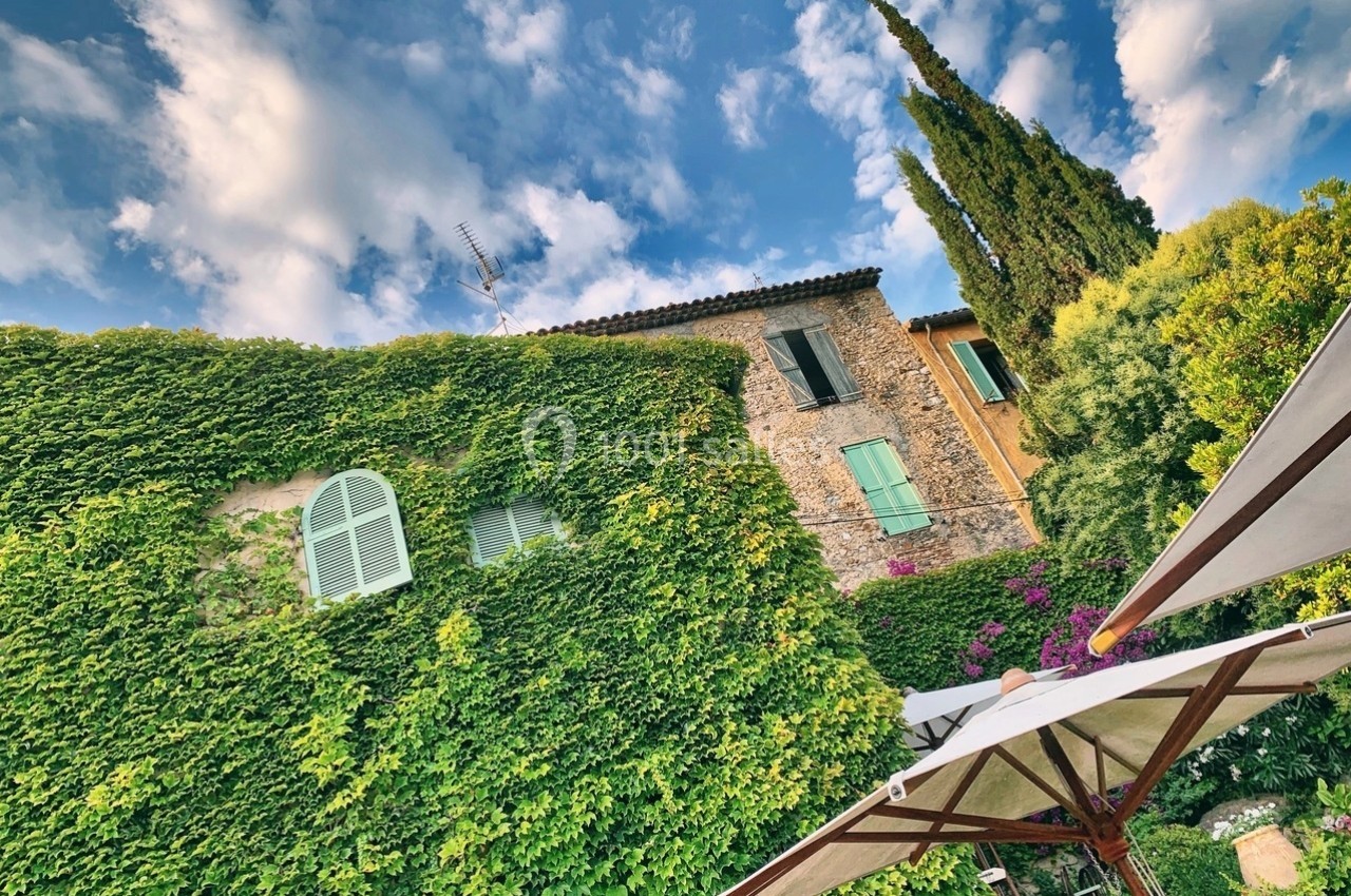 Façade d'une maison en pierre recouverte de lierre, avec volets colorés, arbres et ciel partiellement nuageux.