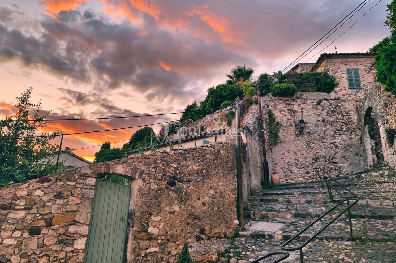 Rue pavée bordée de murs en pierre et végétation, menant à des maisons sous un ciel coloré au coucher du soleil.