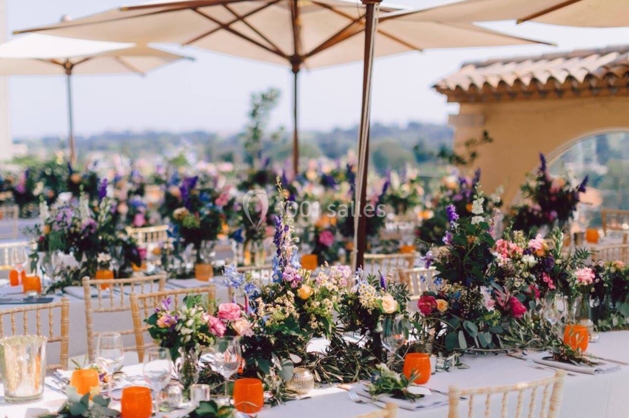 Tables décorées de fleurs colorées et de bougies oranges sous des parasols, en extérieur par temps ensoleillé.
