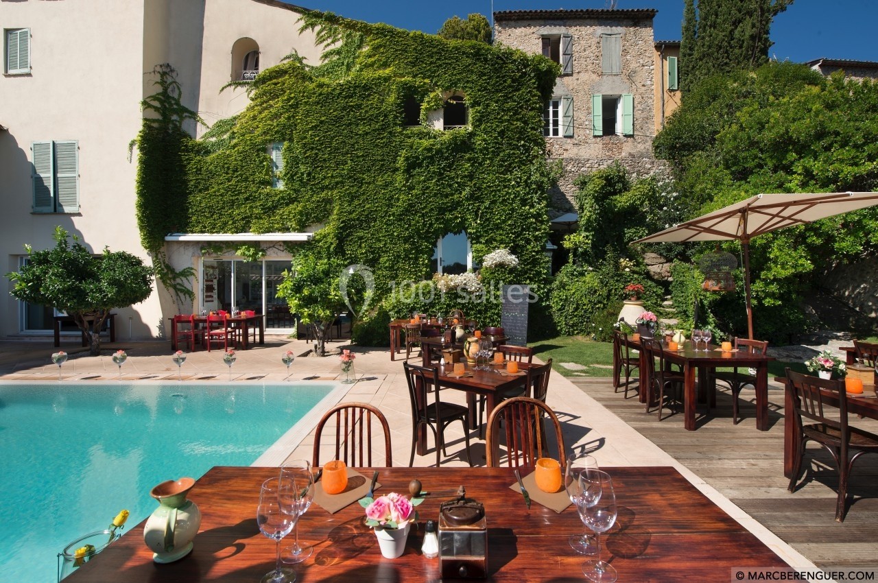 Terrasse avec tables dressées près d'une piscine, entourée de bâtiments couverts de végétation et de verdure.
