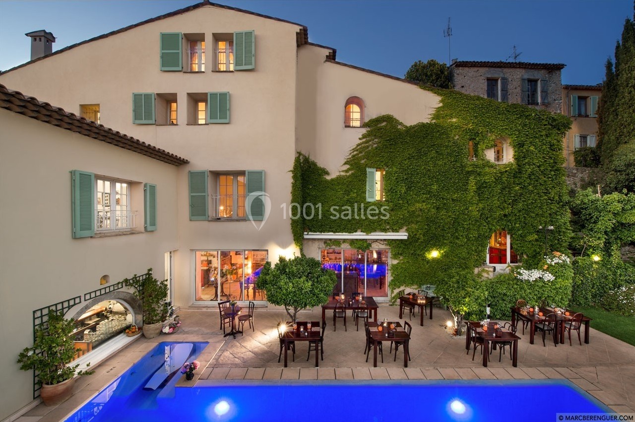 Façade d'une maison avec volets verts, terrasse aménagée, tables dressées et piscine éclairée en soirée.