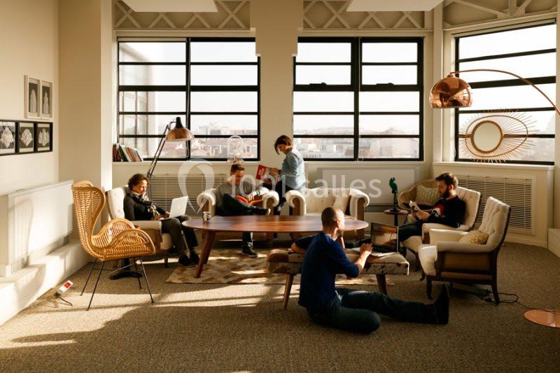 Cinq personnes discutent et lisent dans un salon lumineux avec des fauteuils, une table basse et de grandes fenêtres.