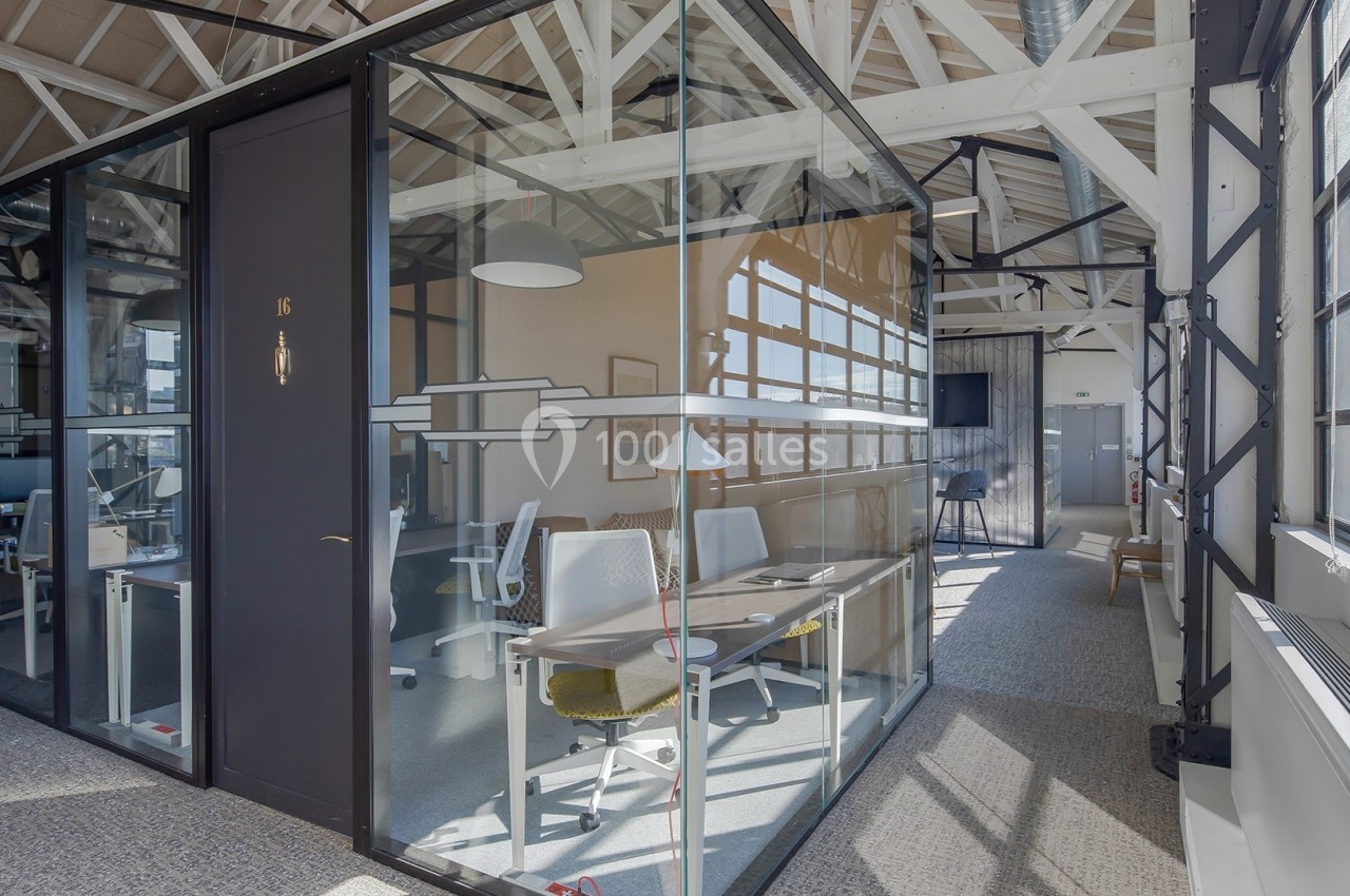 Bureau vitré moderne avec deux postes de travail, situé dans un espace lumineux avec poutres apparentes.