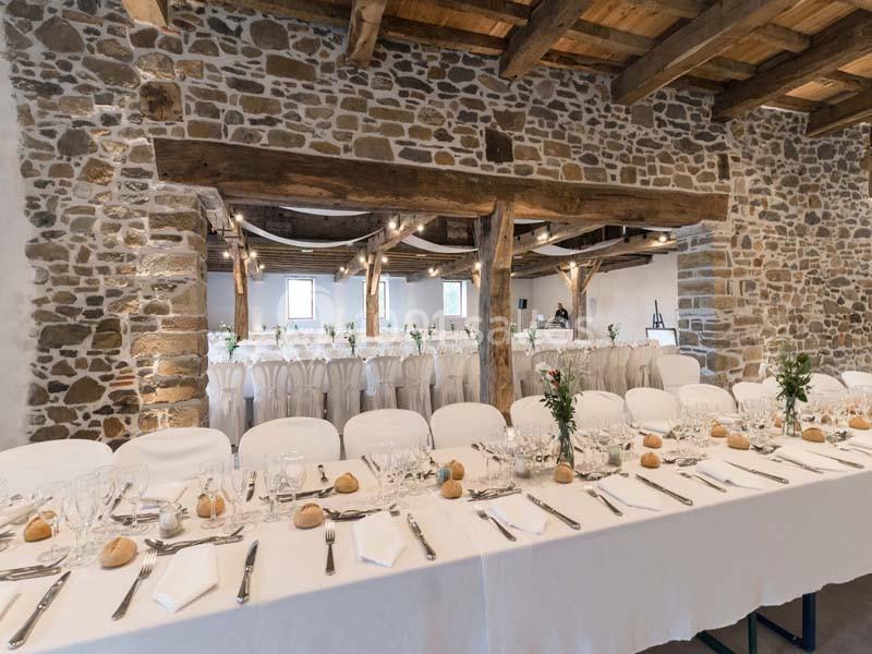 Grande salle de réception en pierre avec tables dressées, nappes blanches, couverts et décorations florales.