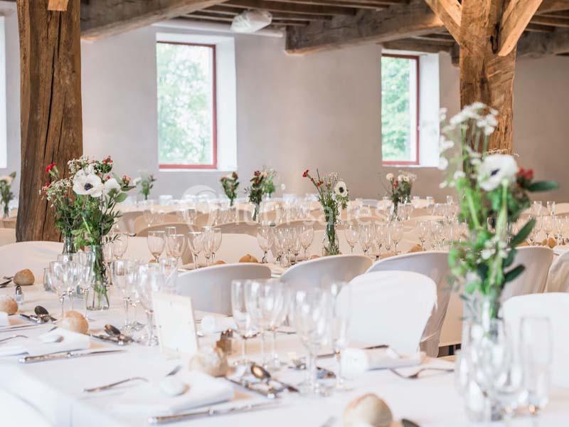 Salle de réception avec tables dressées, décorées de fleurs et de vaisselle, éclairée par des fenêtres.