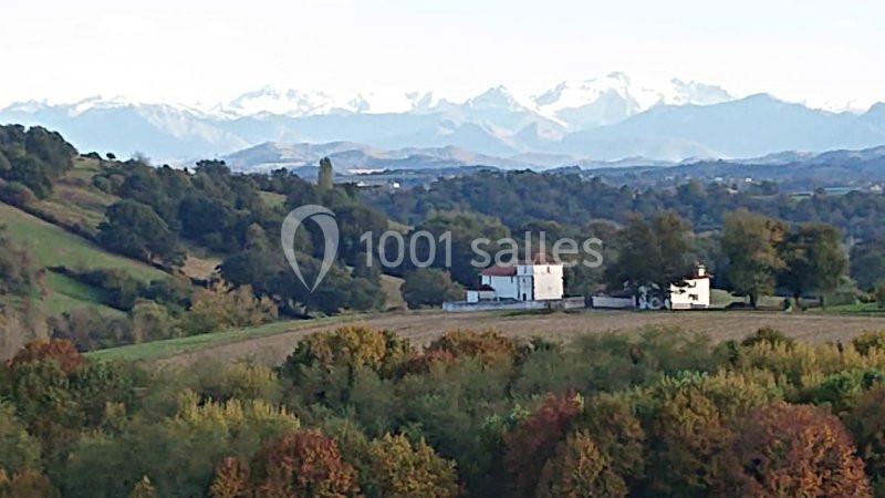 Paysage rural avec des collines verdoyantes, une maison blanche et les montagnes enneigées en arrière-plan.