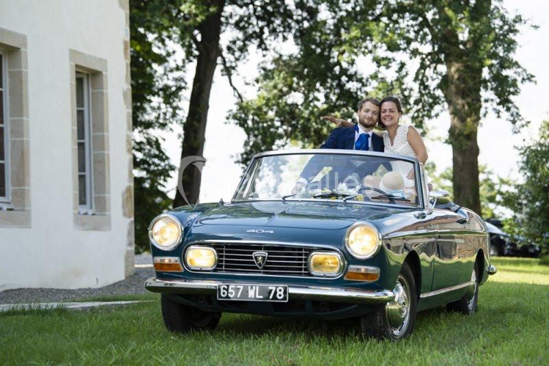 Un couple souriant dans une voiture ancienne décapotable verte, garée sur une pelouse près d'un bâtiment.