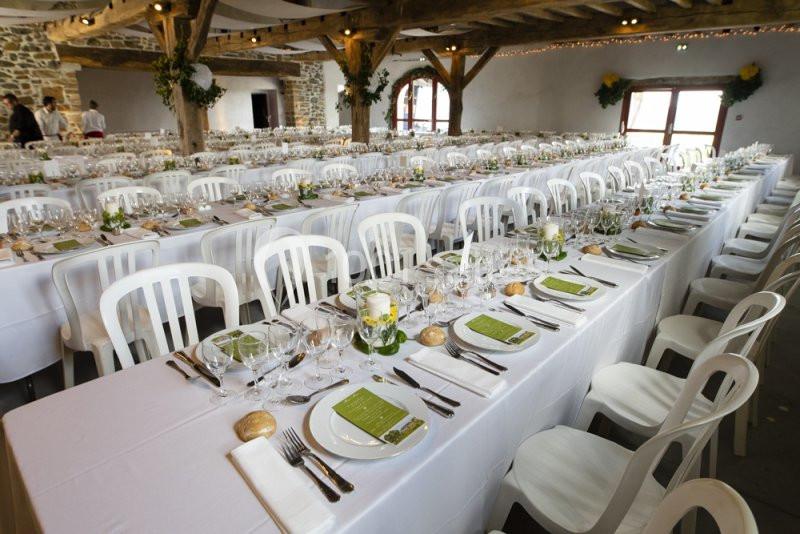Salle de réception avec de longues tables dressées, nappes blanches, couverts et décorations florales.