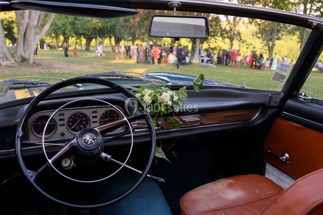 Vue intérieure d'une voiture ancienne avec un volant en métal, des fleurs sur le tableau de bord et un parc en arrière-plan.