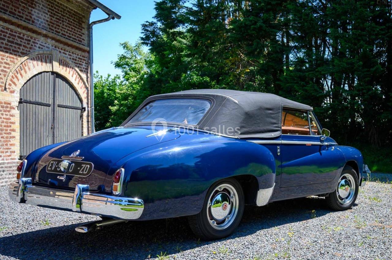 Voiture ancienne bleue avec capote noire, stationnée sur une allée gravillonnée devant un bâtiment en briques.