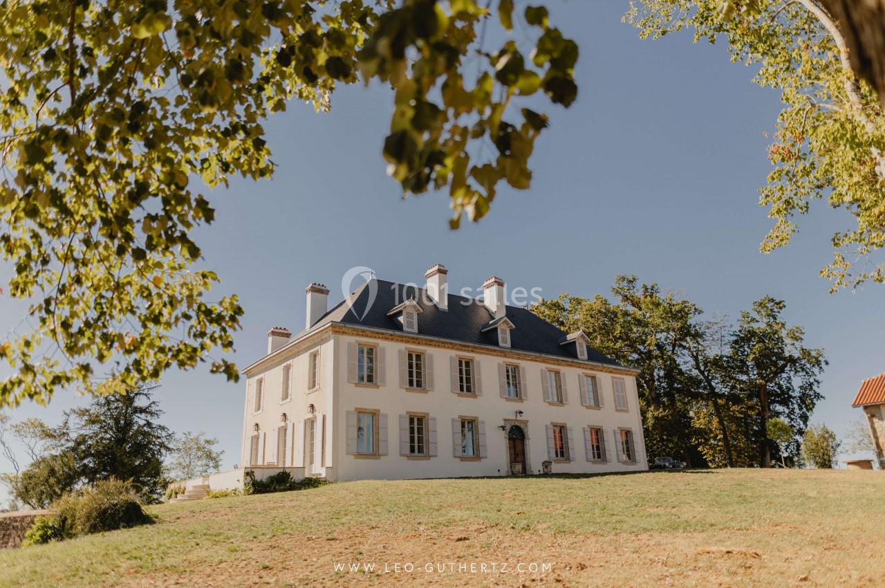 Manoir blanc à deux étages avec toit en ardoise, entouré d'arbres et d'une pelouse sous un ciel dégagé.