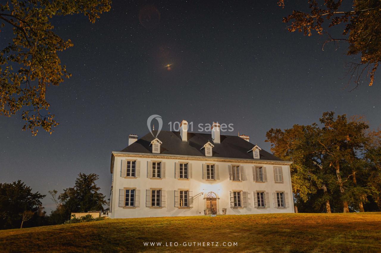 Façade d'un manoir blanc éclairé la nuit, entouré d'arbres sous un ciel étoilé.