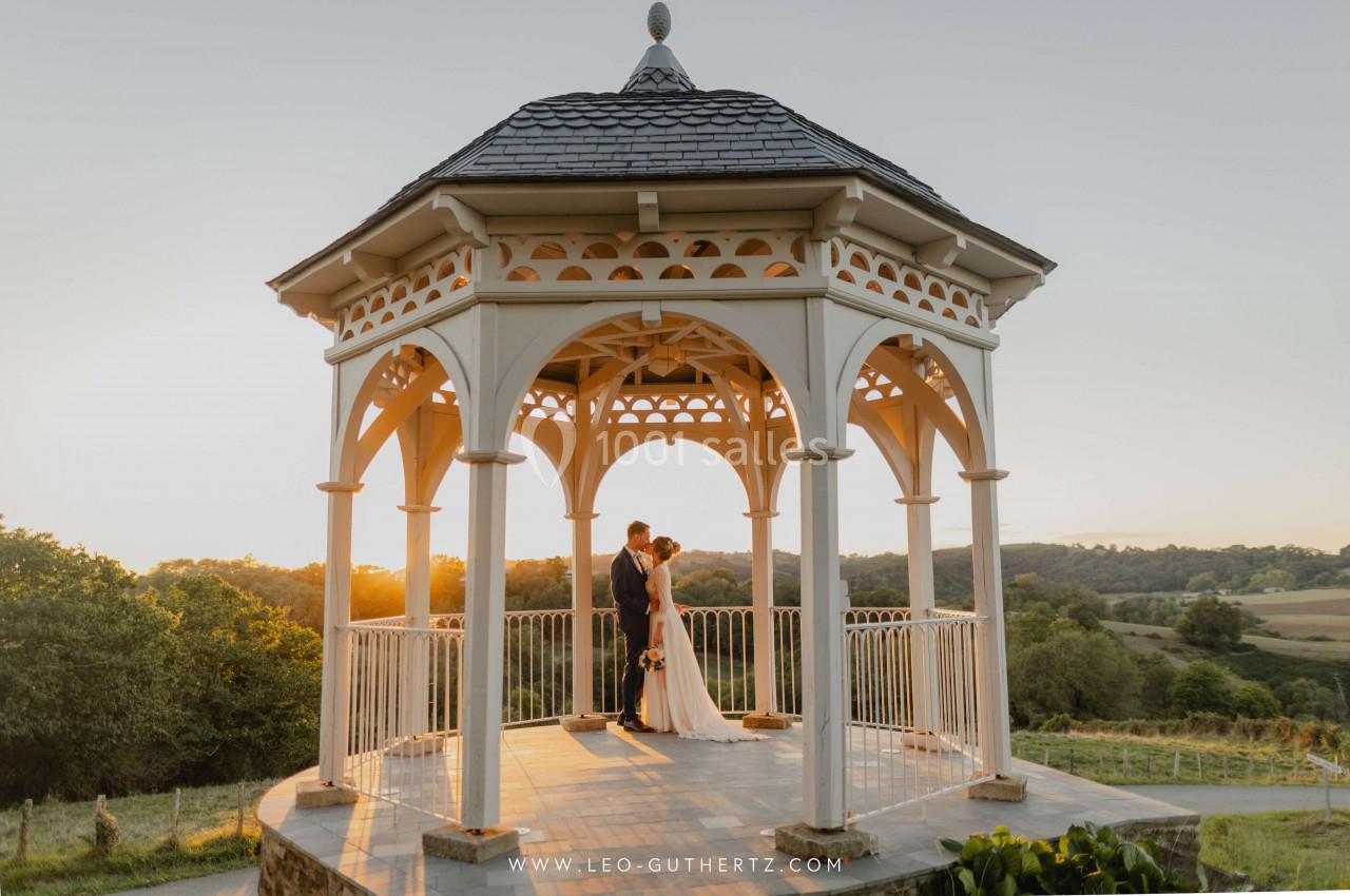 Un couple se tenant sous un kiosque en bois au coucher du soleil, entouré d'un paysage vallonné.