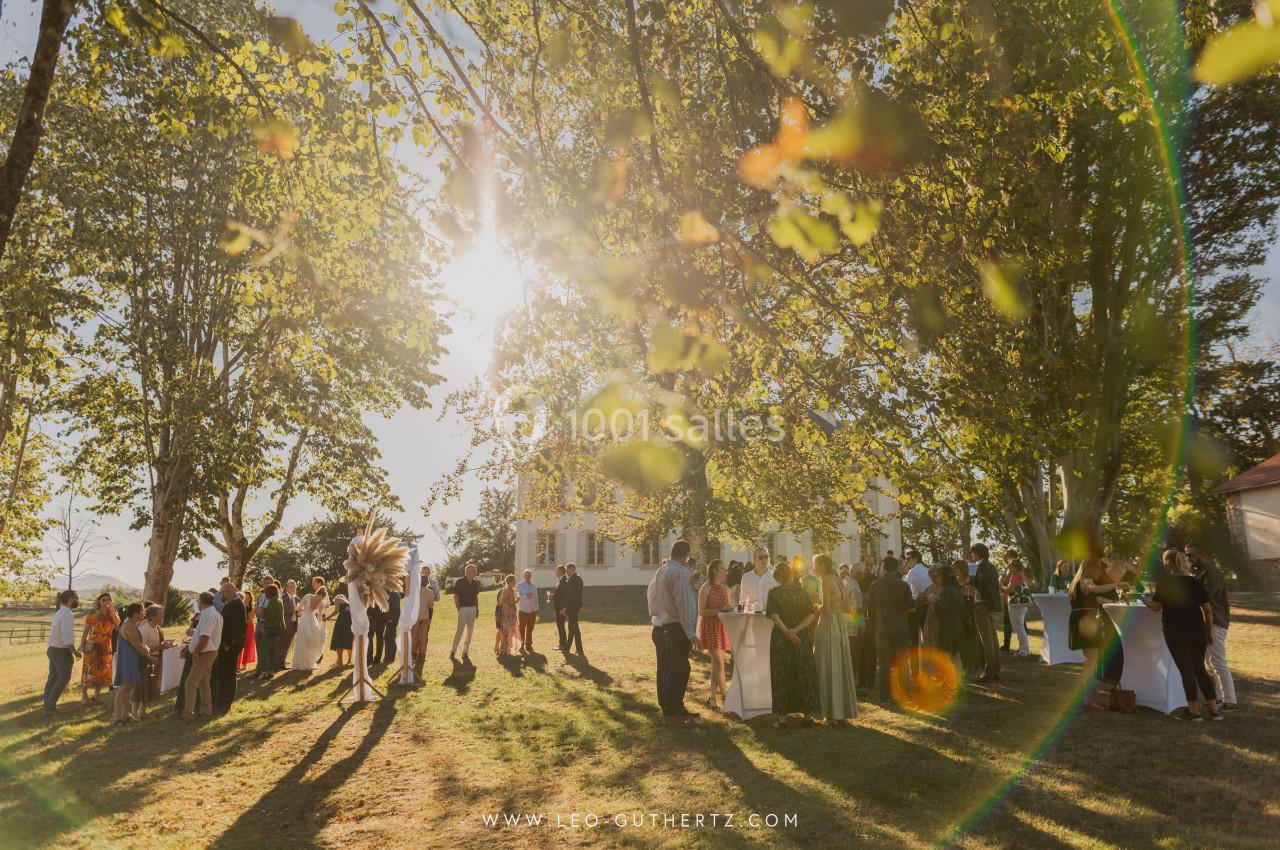 Groupe de personnes rassemblées dans un jardin ensoleillé, entouré d'arbres, lors d'un événement en plein air.